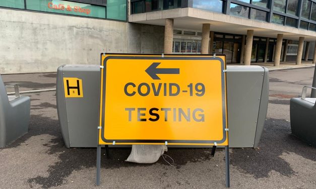 A yellow and black road sign with an arrow pointing left and the words Covid-19 testing