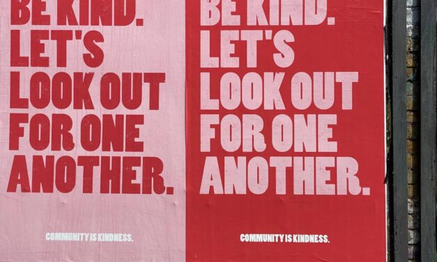 Posters on a billboard, one with red text on a pink background and one with pink on red. Text reads: BE KIND. LET'S LOOK OUT FOR ONE ANOTHER
