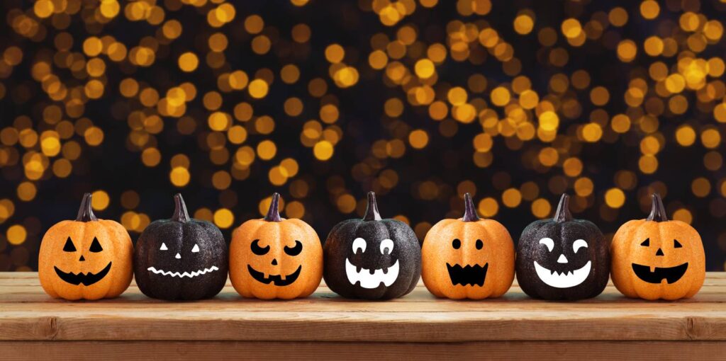 A row of small pumpkins painted with different halloween jack o'lantern faces and alternating black and orange. The background is out of focus orange fairly lights. 