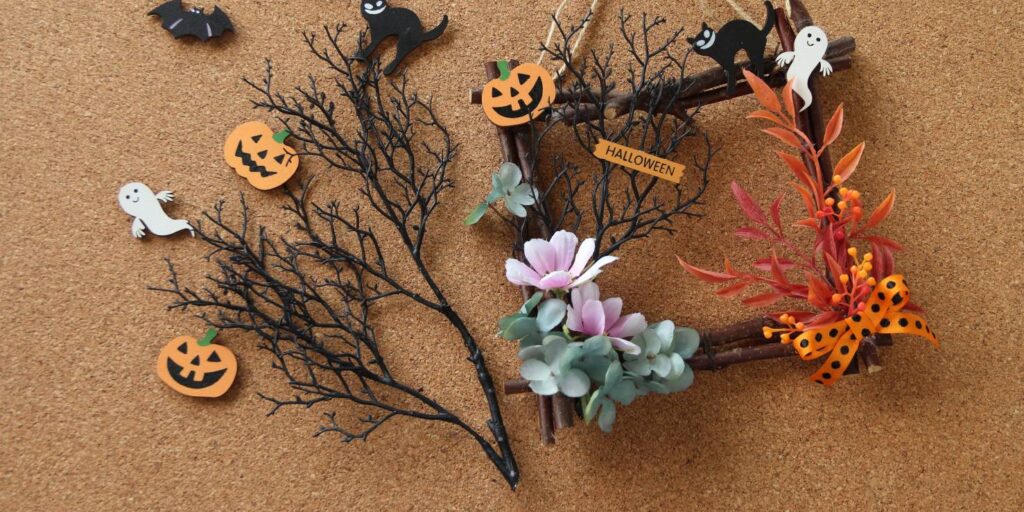 Halloween craft materials making up a wreath of black twigs, orange foliage and pink flowers with cut out shapes of ghosts, cats and pumpkins against a cork background