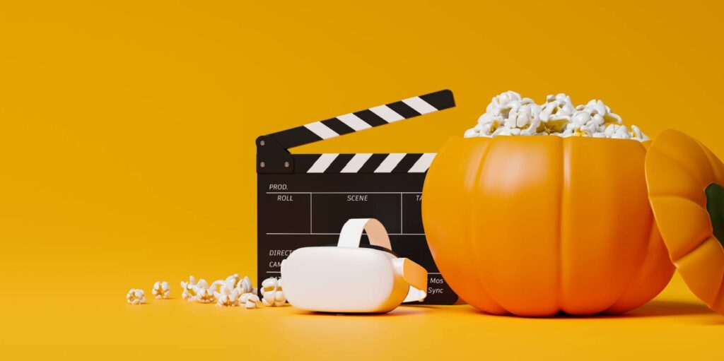 A black and white movie clapper board, white VR headset and orange plastic pumpkin bucket full of popcorn against a bright, plain orange background