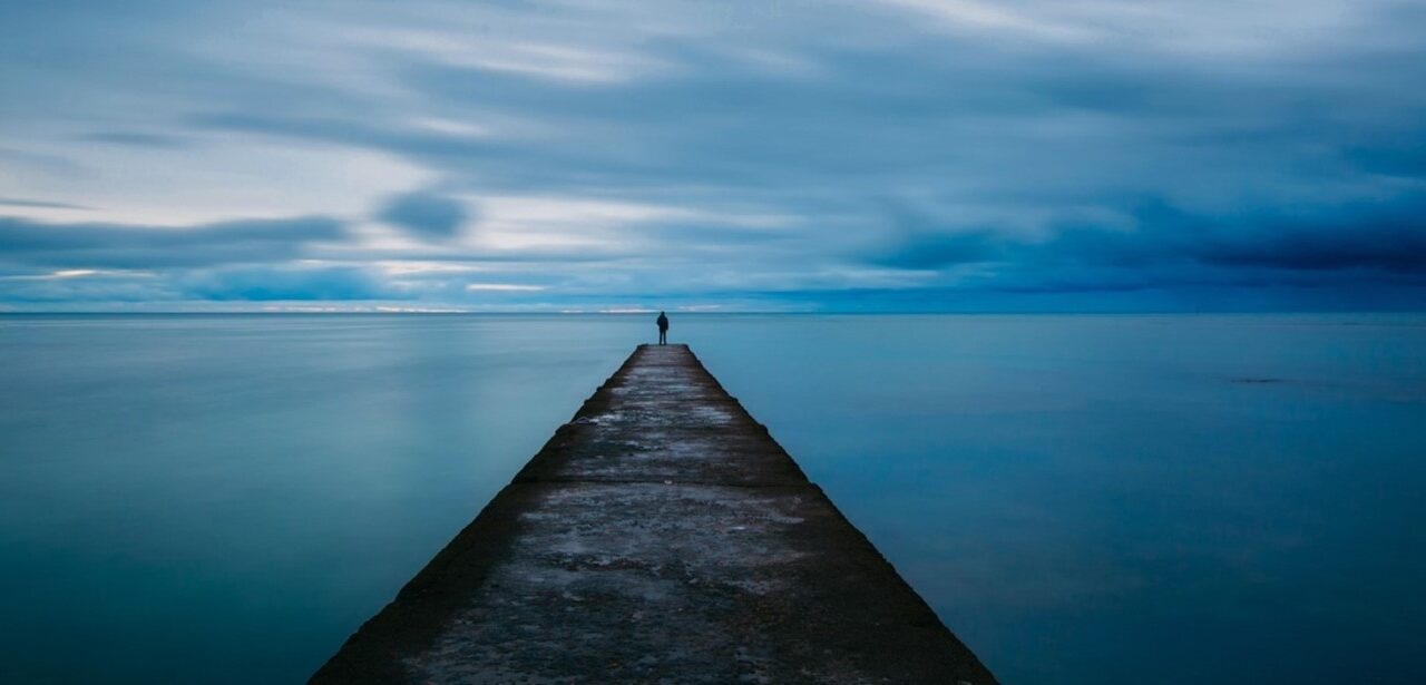 Dark seascape with a concrete jetty leading out. A figure stands at the end, alone.