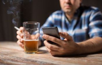 man sits in front of a glass off beer. he is smoking and looking at his phone