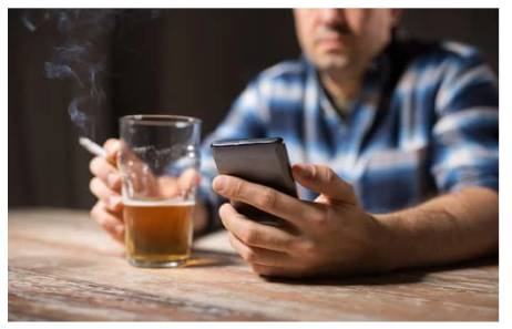 man sits in front of a glass off beer. he is smoking and looking at his phone