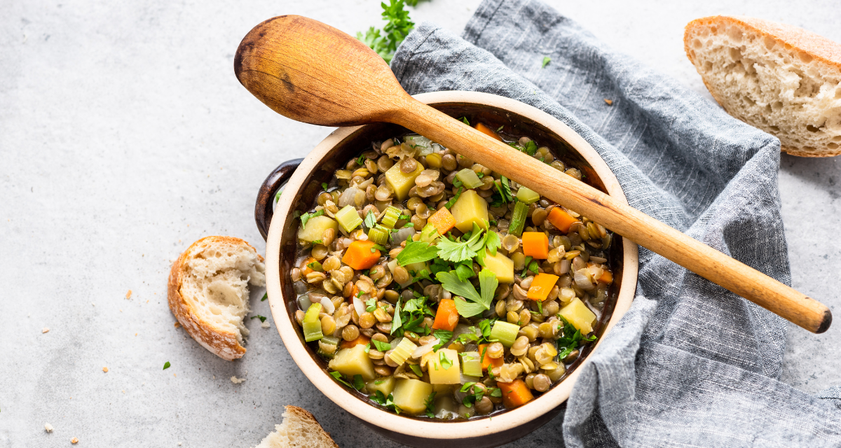 A casarole dish full with a vegetarian type stew sits on top of a light blue tea towel. A wooden sppon lays across the top of the dish and crusty bread sits at the side | World vegetarian day recipes.