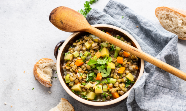 A casarole dish full with a vegetarian type stew sits on top of a light blue tea towel. A wooden sppon lays across the top of the dish and crusty bread sits at the side | World vegetarian day recipes.