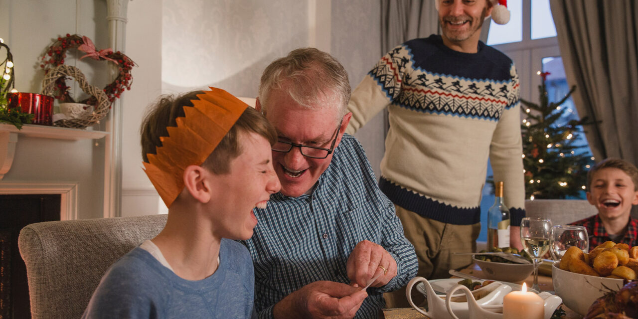 A man and a boy sitting at a festive table laughing over a cracker joke at Christmas