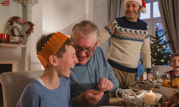 A man and a boy sitting at a festive table laughing over a cracker joke at Christmas