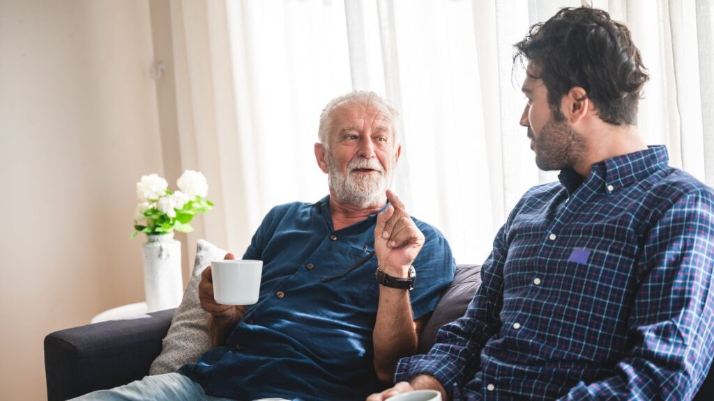 An older man, holding a cup of tea, talks to a younger man in a sitting room setting
