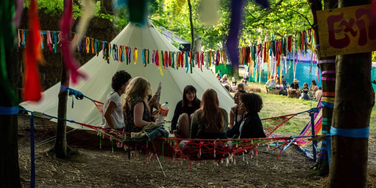a group of women sitting on the ground under trees in front of a teepee style tent draped with bunting