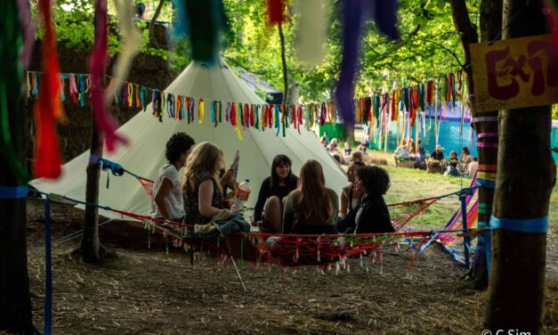 a group of women sitting on the ground under trees in front of a teepee style tent draped with bunting