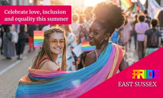 two people stand in a crowd shoulder to shoulder smiling at the camera. A rainbow flag is draped across their shoulders