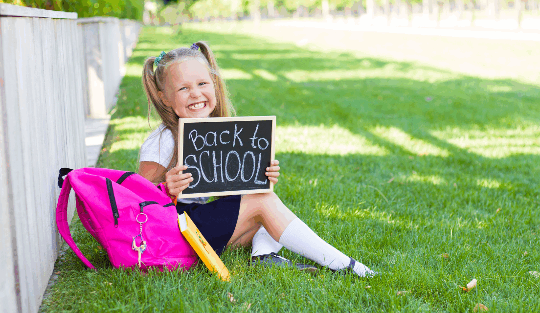 A young girl in school uniform sits on the grass with a pink backpack holds a board that reads East Sussex back to school.