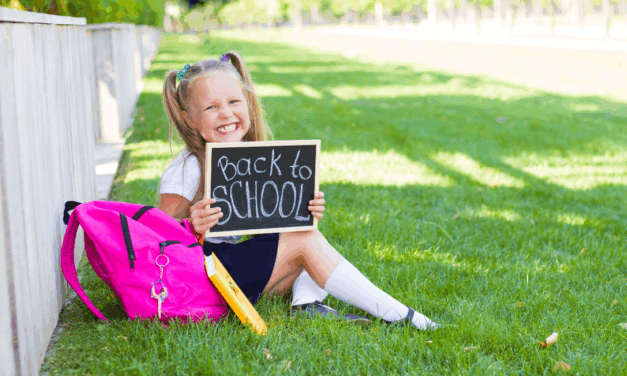 A young girl in school uniform sits on the grass with a pink backpack holds a board that reads East Sussex back to school.
