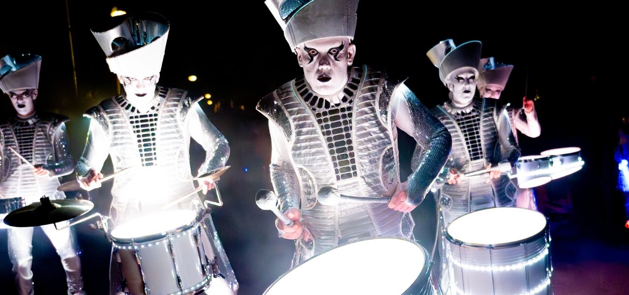 four drummers in silver costumes lit from below
