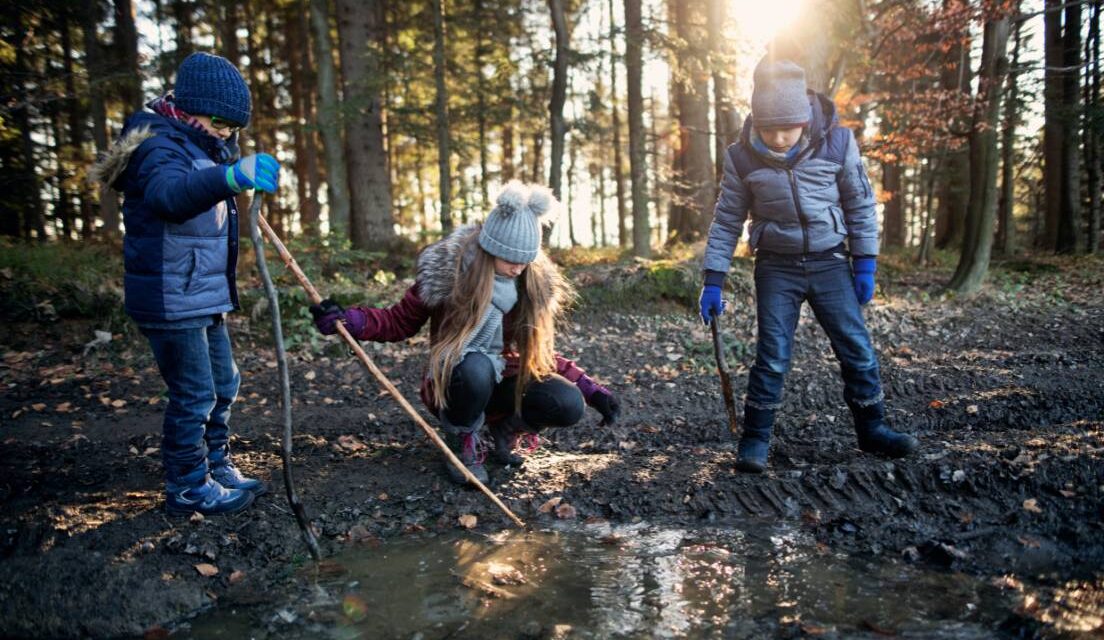 Three children in a forest dressed in warm hats and coats, playing with sticks in a puddle as they enjoy half term in East Sussex.