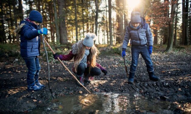 Three children in a forest dressed in warm hats and coats, playing with sticks in a puddle as they enjoy half term in East Sussex.