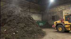 A large pile of compost in a warehouse. A yellow tractor is nearby. 
